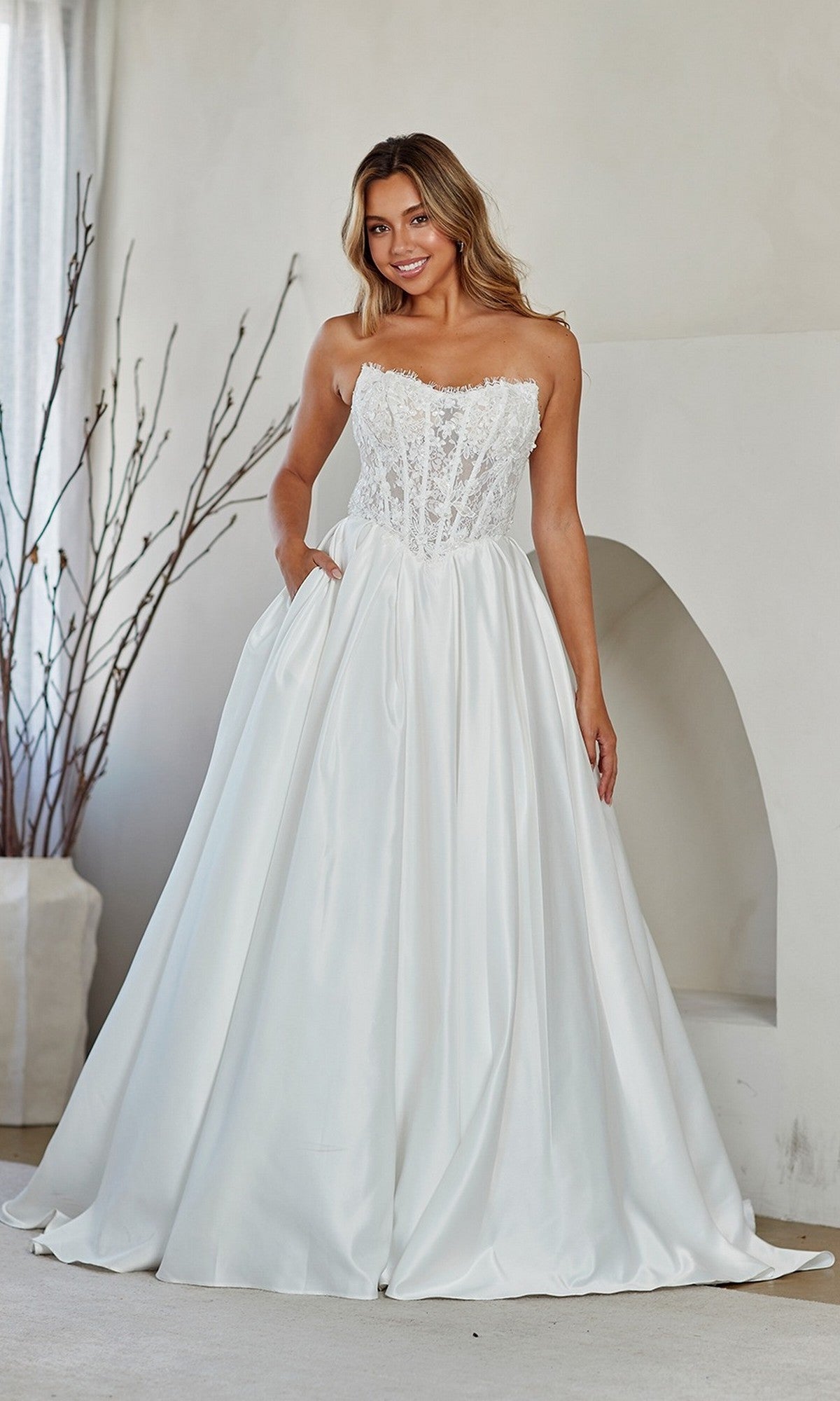 A woman stands indoors wearing the Amelia Couture 2089, a long white prom dress featuring a strapless lace corset bodice and full satin skirt, echoing elegant A-line bridal style.